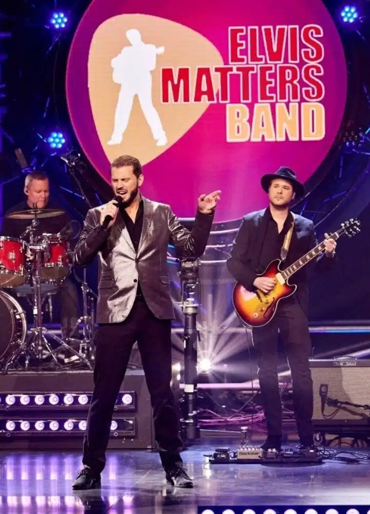 Zanger van de Elvis Matters Band in zilveren colbert zingt op het podium, met gitarist en drummer